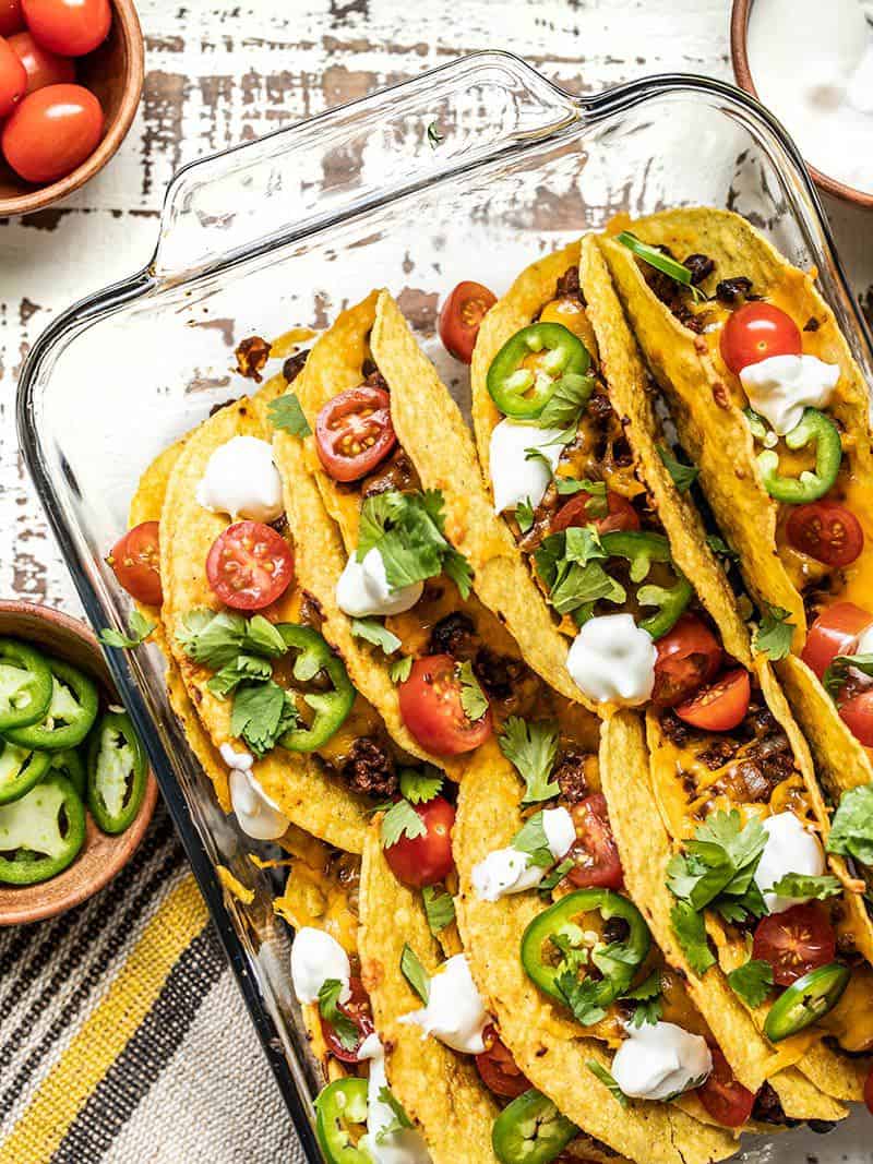 A glass casserole dish filled with Baked Beef and Black Bean Tacos with bowls of toppings on the sides 