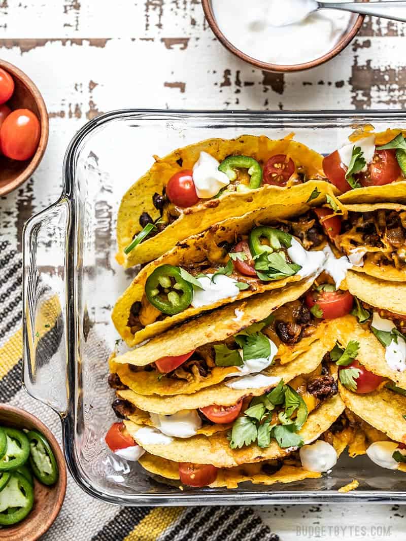 A glass casserole dish filled with baked beef and black bean tacos, with bowls of toppings on the sides 