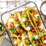 A glass casserole dish filled with baked beef and black bean tacos, with toppings in bowls on the sides