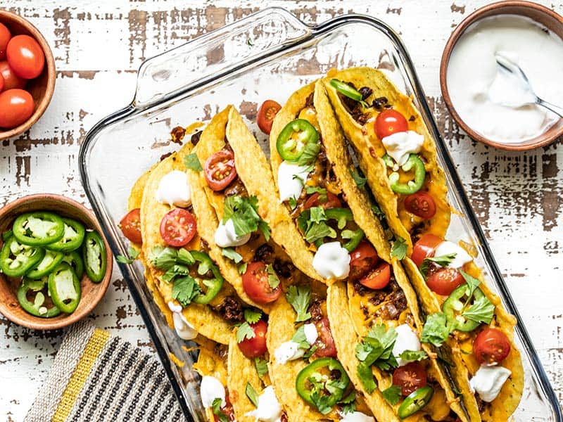 A glass casserole dish filled with baked beef and black bean tacos, with toppings in bowls on the sides