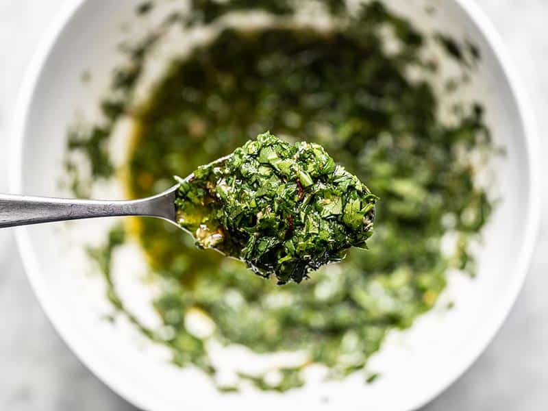 Close up of a spoonful of chimichurri sauce, the bowl in the background.