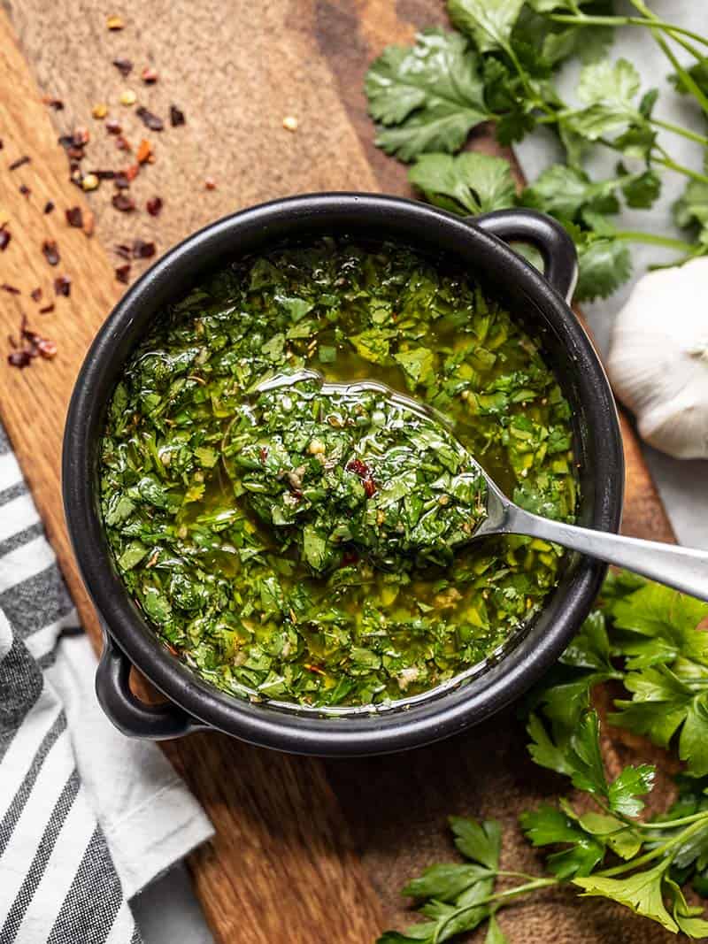 A black ceramic bowl filled with fresh chimichurri sauce on a wooden board, with crushed peppers, garlic, and cilantro on the sides