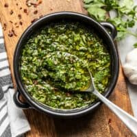 A black ceramic bowl of chimichurri with a spoon, sitting on a wooden cutting board.