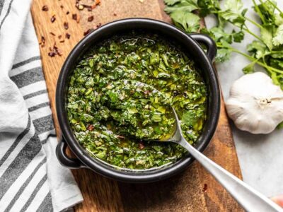 A black ceramic bowl of chimichurri with a spoon, sitting on a wooden cutting board.