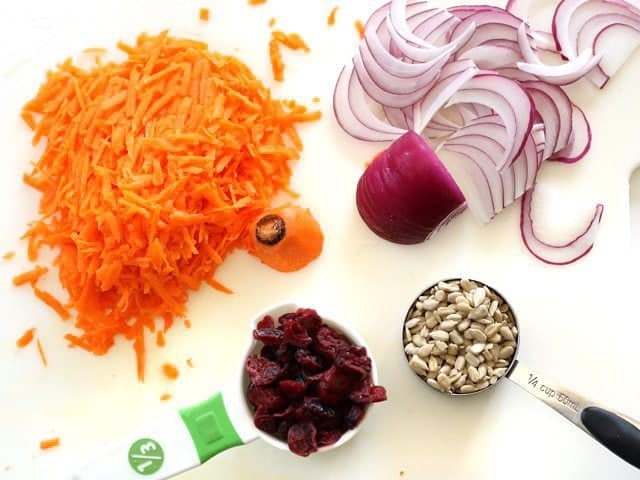 Shredded carrots, sliced red onion, sunflower seeds, and dried cranberries on a cutting board