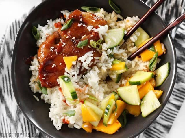 Sticky Ginger Soy Glazed Chicken in a bowl with jasmine rice and cucumber mango salad, being eaten with chopsticks