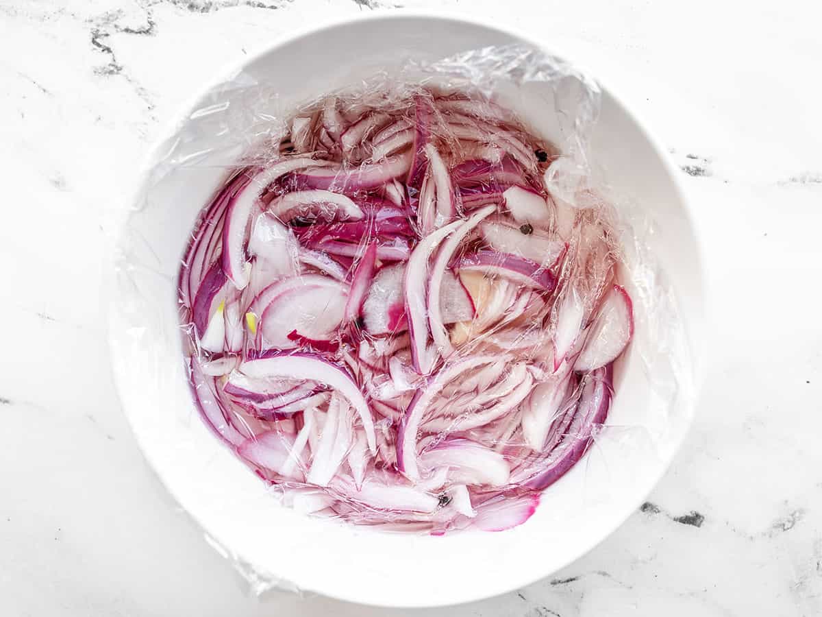 Vinegar in a bowl with sliced onions, plastic wrap on top