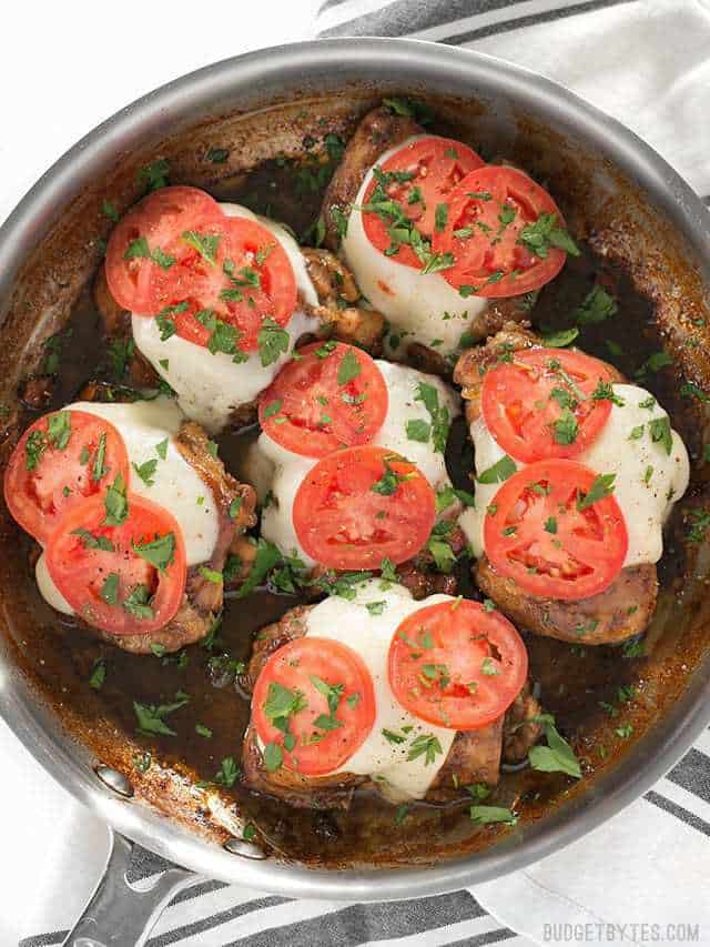 Overhead view of a skillet full of balsamic chicken thighs with melted mozzarella, fresh tomatoes, and chopped parsley on top.
