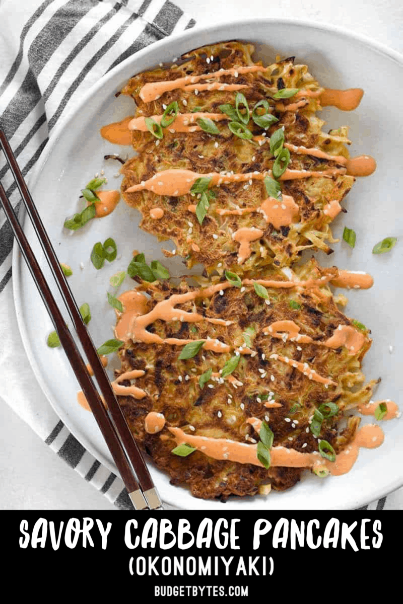 Two savory cabbage pancakes on a plate with sriracha mayo, green onion, sesame seeds, and chopsticks on the side.