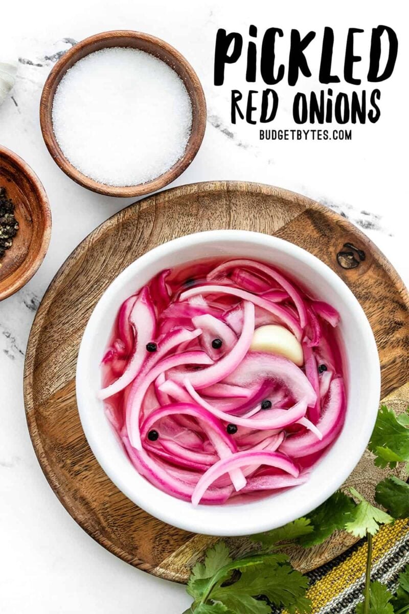 Overhead view of a bowl full of pickled red onions on a wooden plate with cilantro and salt on the side