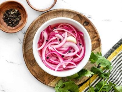 Overhead view of pickled red onions in a bowl on a wooden plate next to bowls of peppercorns and salt.