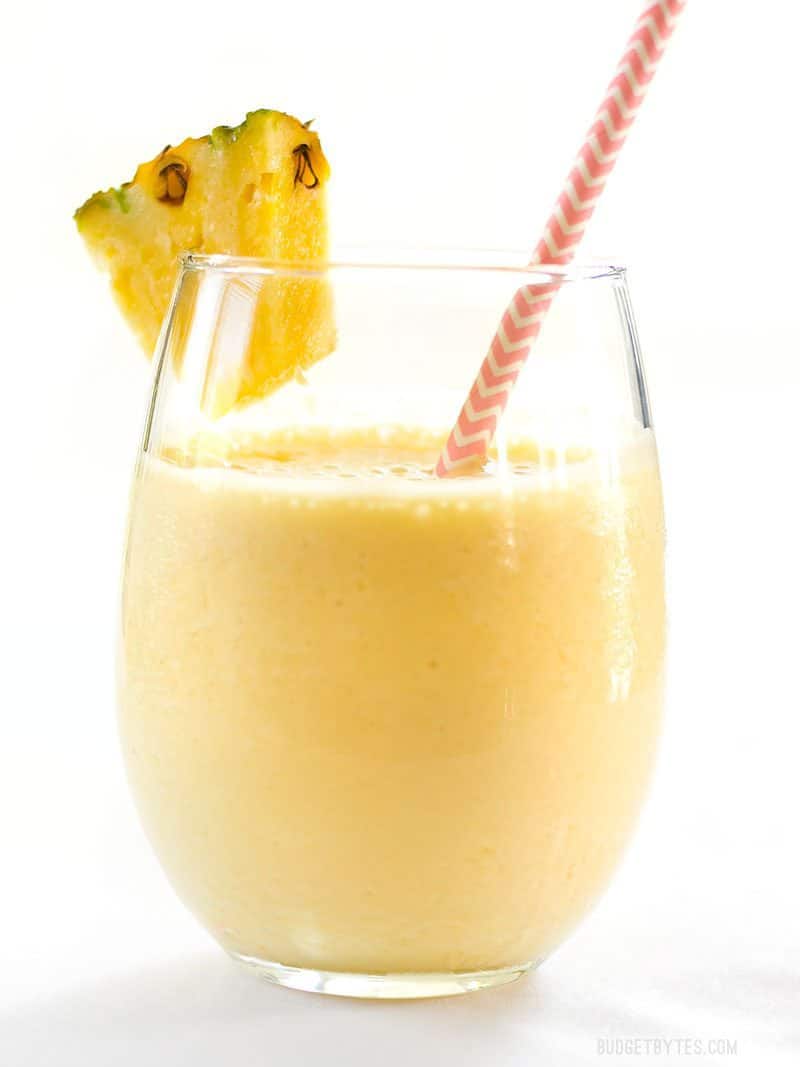 A round glass with Pineapple Orange Julius, a pink and white stripped paper straw, and a slice of pineapple on the rim.