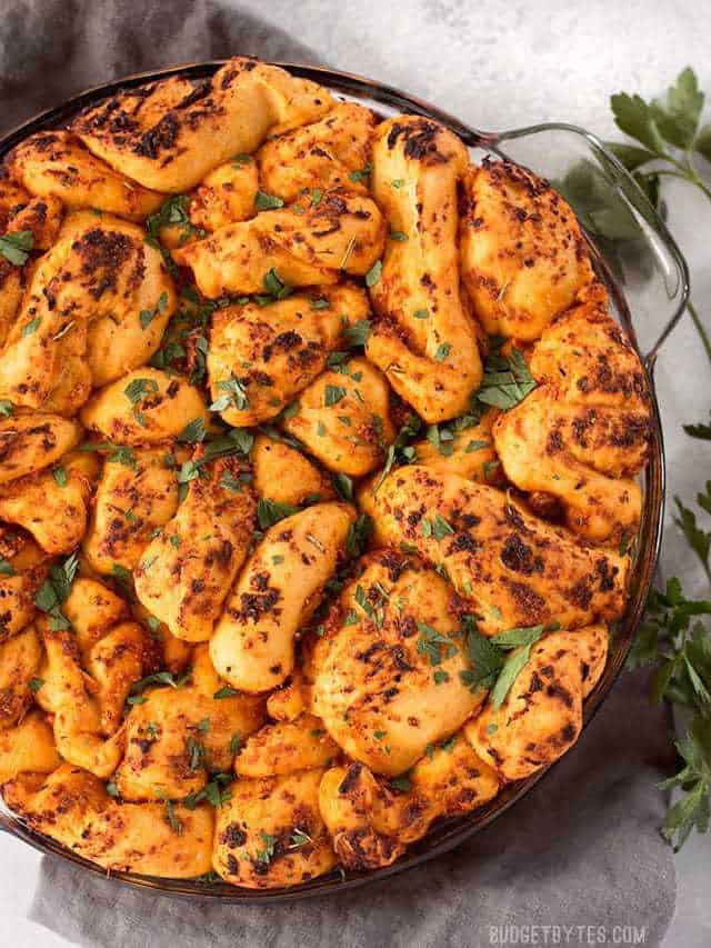 Tomato Herb Pull Apart Bread combines a rich and tangy tomato sauce, savory Parmesan, and fresh bread dough for an irresistible appetizer loaf. BudgetBytes.com Overhead view of a round glass baking dish filled with Tomato Herb Pull Apart Bread, parsley garnish on the side