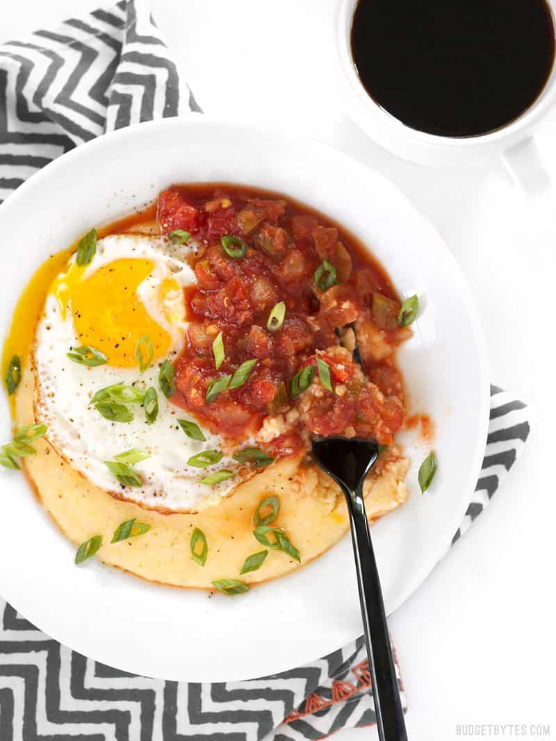 Cheddar Grits Breakfast Bowls are an easy but indulgent breakfast or brunch treat that you can whip up in no time. BudgetBytes.com A Cheddar Grits Breakfast Bowl on a striped napkin, being eaten with a black fork