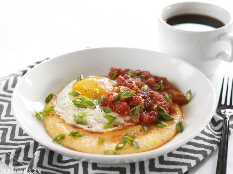 Cheddar Grits Breakfast Bowls are an easy but indulgent breakfast or brunch treat that you can whip up in no time. BudgetBytes.com Front view of a Cheddar Grits Breakfast Bowl with a cup of coffee behind it