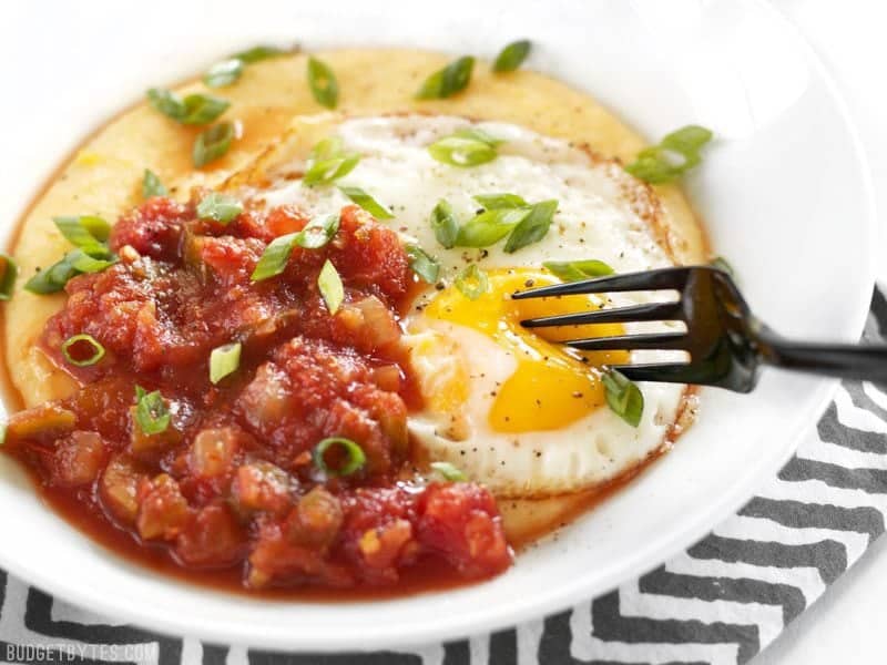 Cheddar Grits Breakfast Bowls are an easy but indulgent breakfast or brunch treat that you can whip up in no time. BudgetBytes.com The yolk of an egg in a Cheddar Grits Breakfast Bowl being pierced with a fork.