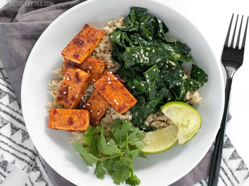 A Chili Garlic Tofu Bowl garnished with lime wedges and cilantro, a fork on the side.