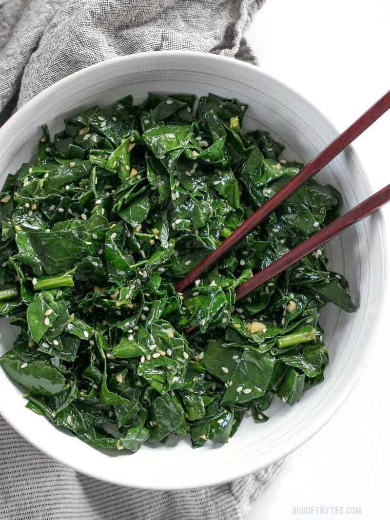 A big bowl of Sesame Kale with wooden chopsticks about to pick up a piece.