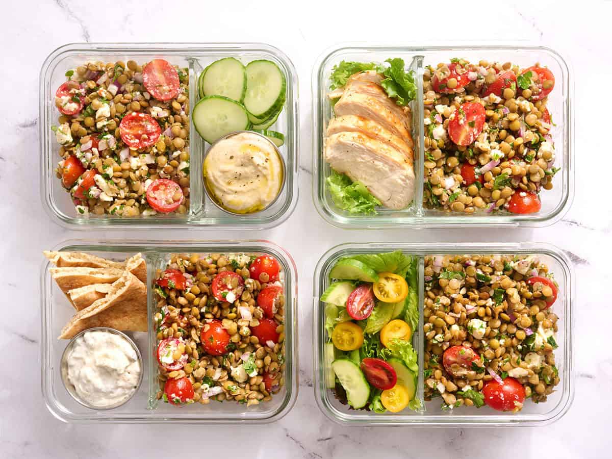 Overhead view of four meal prep containers with lentil salad, tzatziki sauce, cucumber slices and grilled chicken.