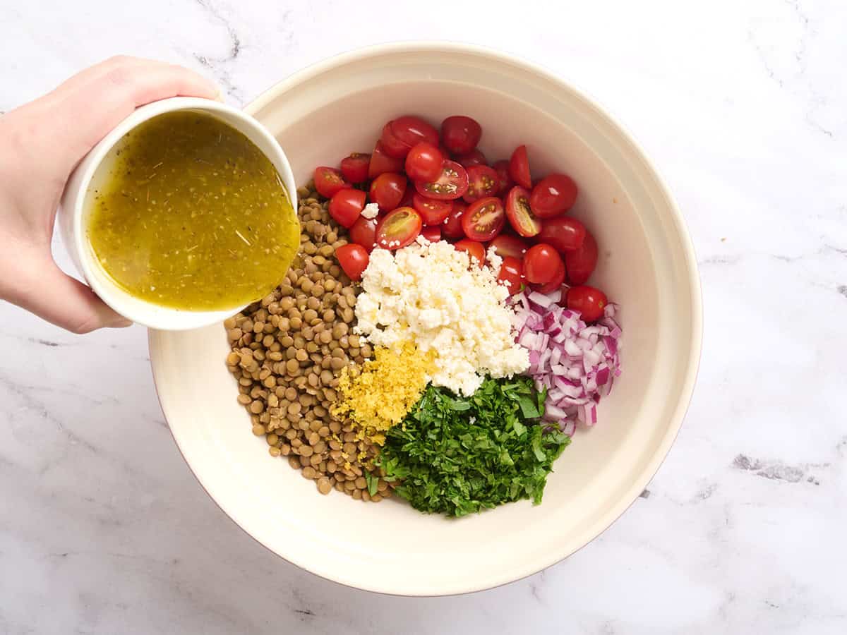 Dressing being poured into a bowl with tomatoes, feta, parsleyy, red onion and cooked lentils.