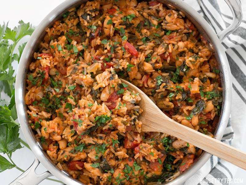 Overhead view of Tomato Herb Rice with White Beans and Spinach being scooped out of the skillet with a wooden spoon.