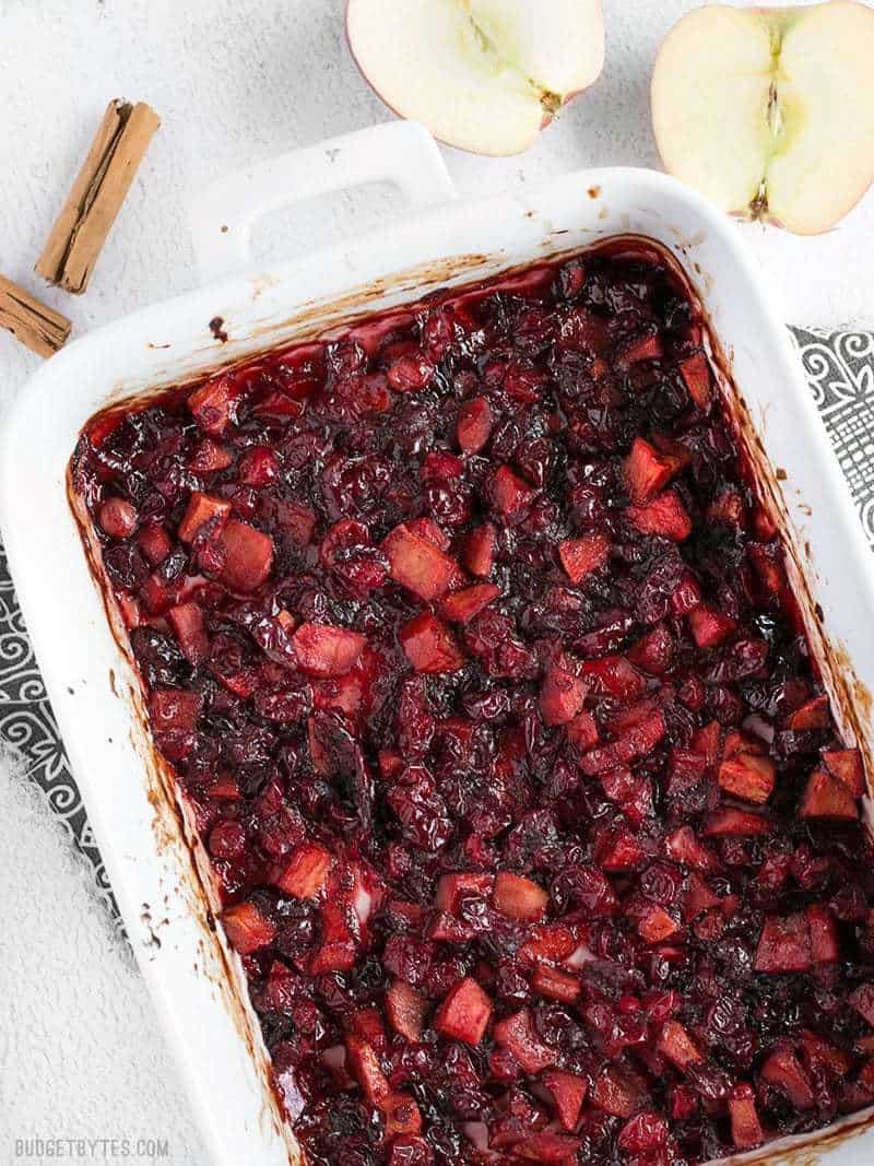 A white casserole dish full of Roasted Apple Cranberry Relish with cinnamon sticks and apples on the sides.