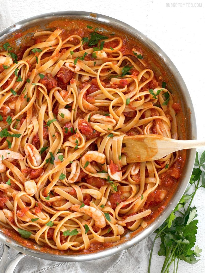 A skillet full of Spicy Seafood Pasta with Tomato Butter Sauce with a wooden fork twirled in the pasta and parsley on the side.