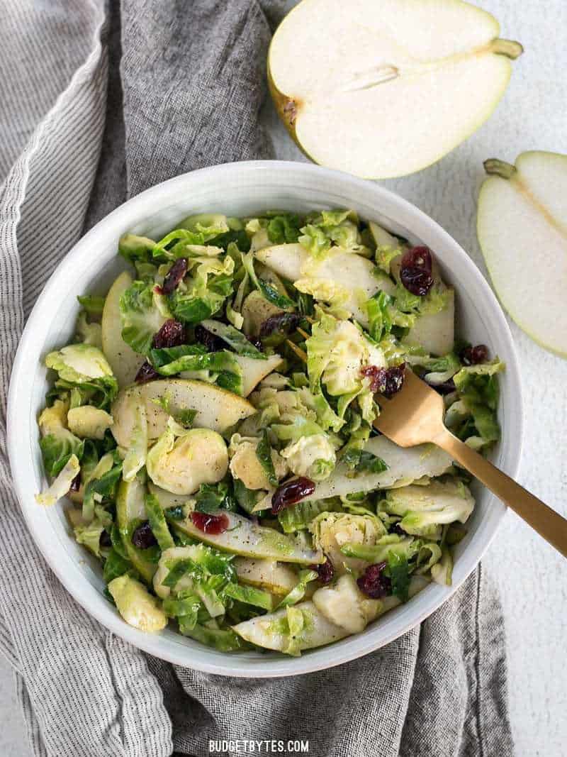 A bowl of Warm Brussels Sprouts and Pear Salad with a gold fork, next to a fresh pear cut in half.