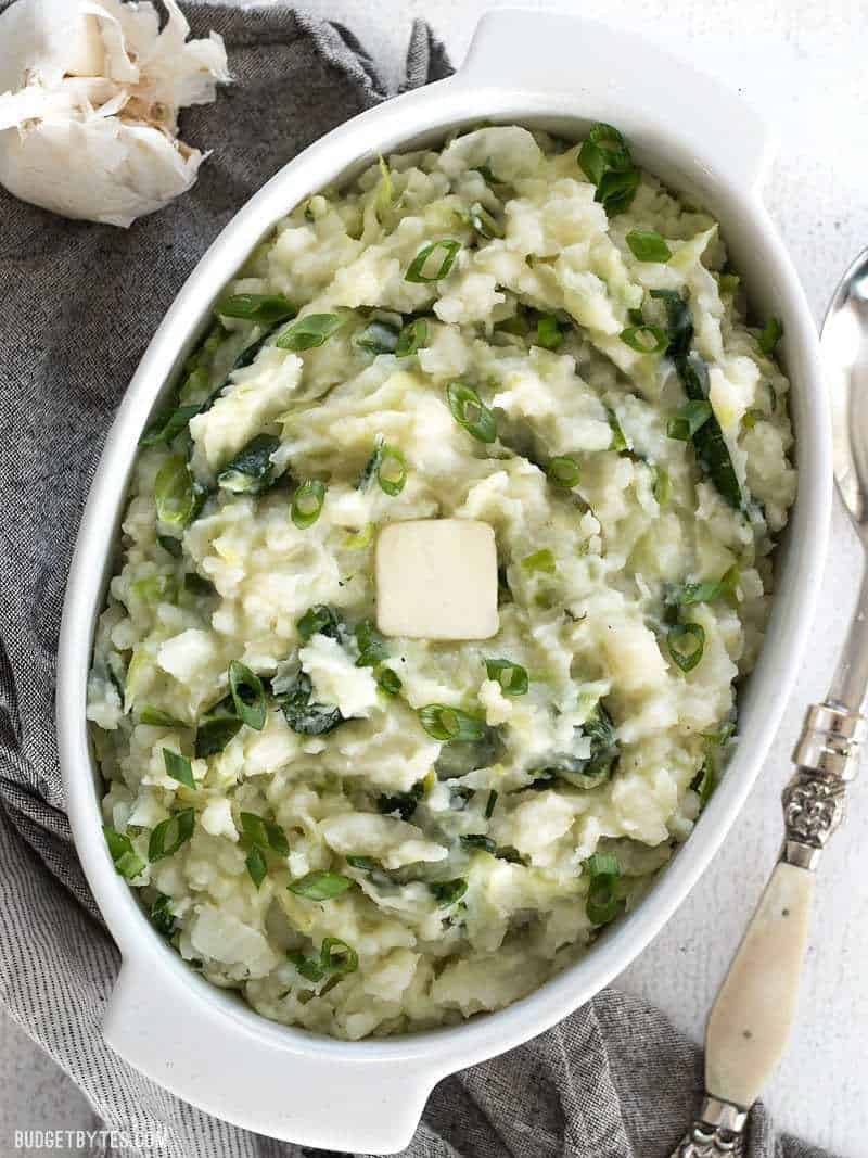 A large casserole dish full of Colcannon with a pat of butter melting on top and a head of garlic sitting on the side