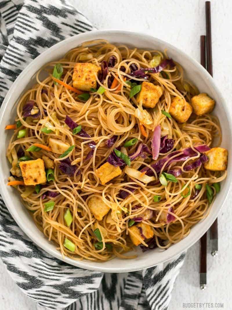 A big bowl of Singapore Noodles with Crispy Tofu on a patterned black and white napkin with chopsticks on the side