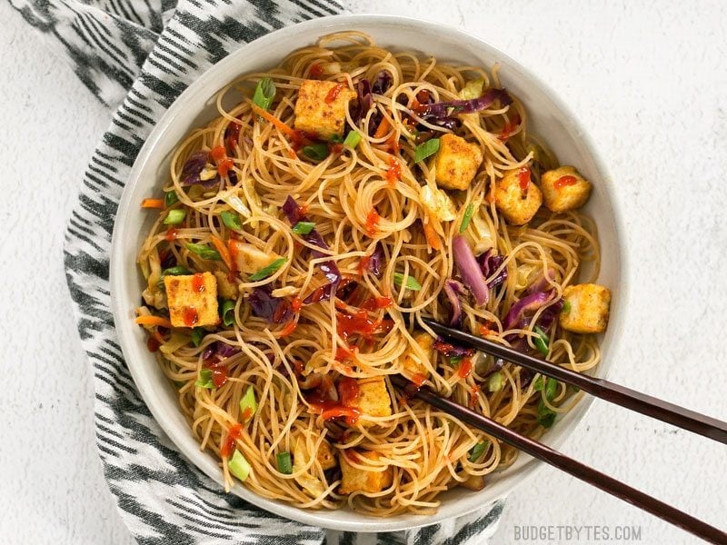 Overhead view of Singapore Noodles with Crispy Tofu garnished with sriracha, chopsticks in the bowl