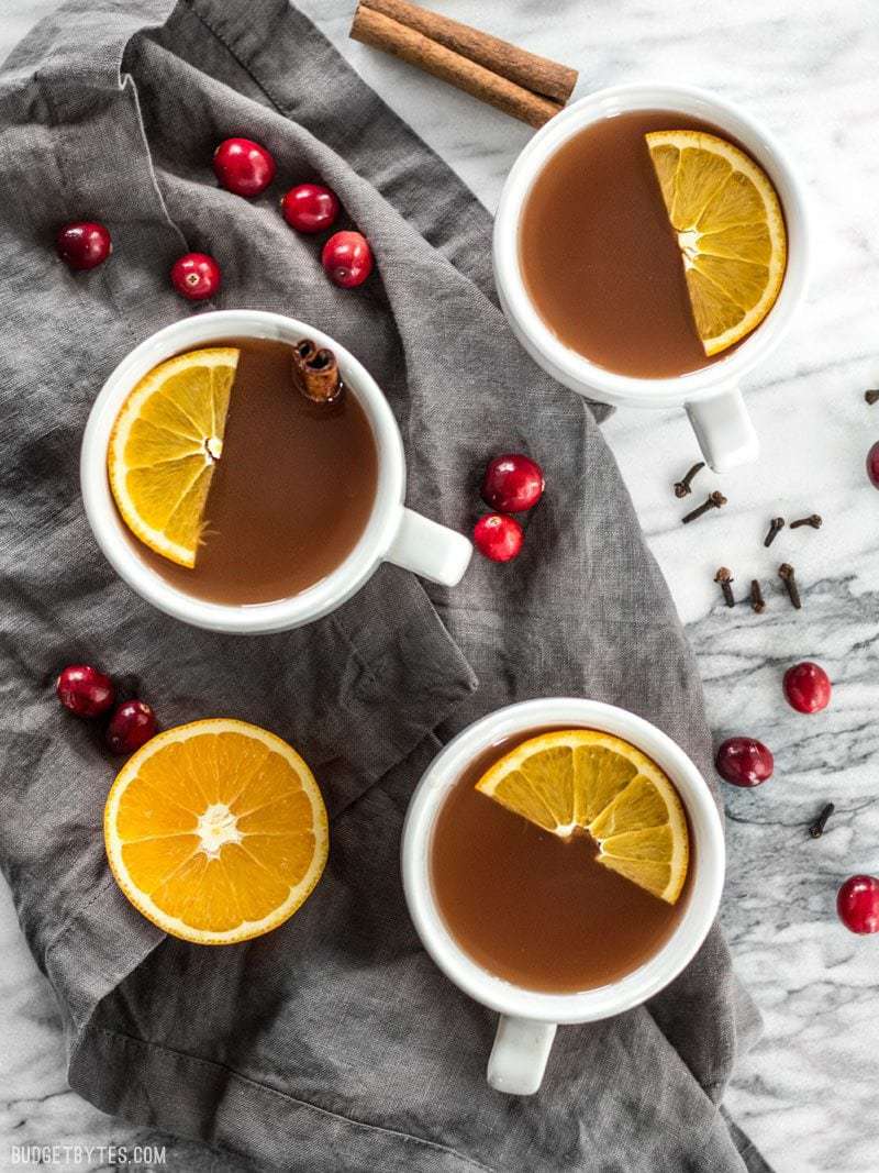 Three mugs of Slow Cooker Spiced Cranberry Apple Cider with fresh orange slices floating on top, and cranberries, cinnamon, and cloves sprinkled around