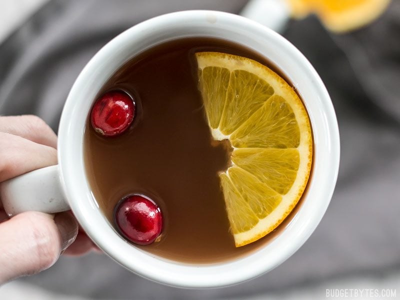 Slow Cooker Spiced Cranberry Apple Cider is an easy and festive drink for all your holiday party guests. BudgetBytes.com Close up of a mug full of Slow Cooker Spiced Cranberry Apple Cider with cranberries and an orange slice floating on top