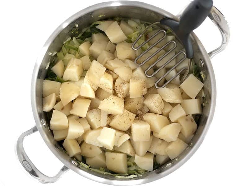 Add Potatoes, Peppe, and Milk to stock pot with cabbage. A potato masher in the pot, ready to mash.