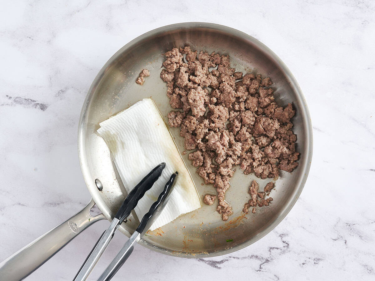 A paper towel mopping up grease from a skillet with ground beef.