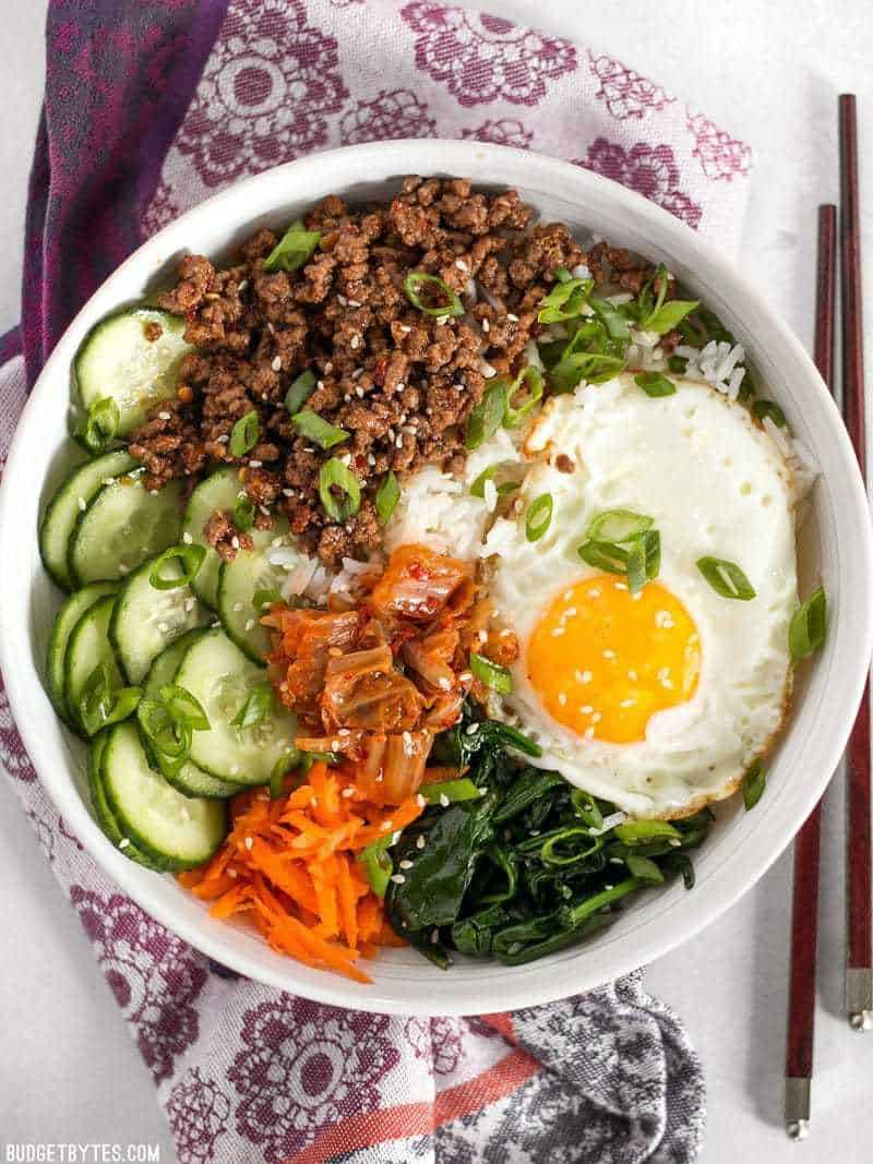A colorful bowl of Bibimbap with an over easy egg, sitting on a purple floral napkin.