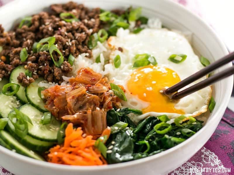 Side view of a bowl of Bibimbap with the yolk being broken by a pair of chopsticks.