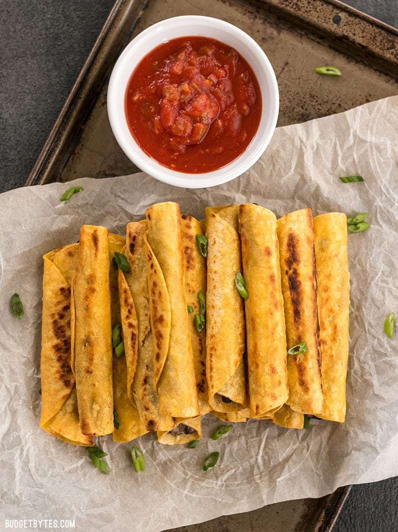 A pile of Creamy Black Bean Taquitos on a piece of parchment with a dish of salsa on the side