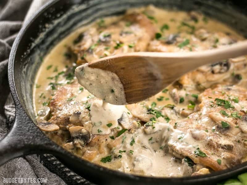 Creamy Garlic sauce dripping off a wooden spoon onto the chicken thighs in the skillet