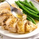 A spoon drizzling jus on a herb roasted chicken breast with green beans.