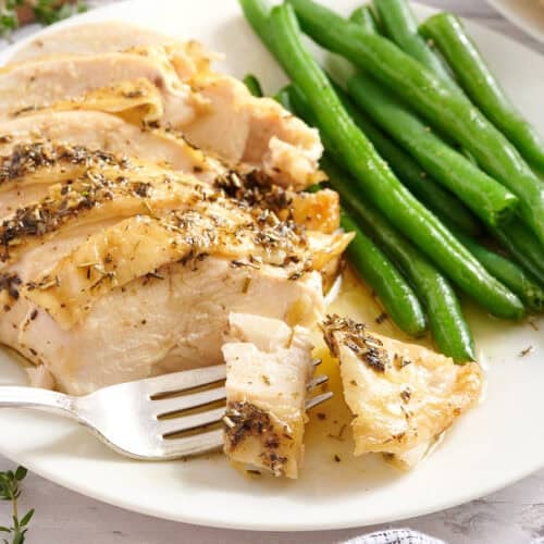 A herb roasted chicken breast with green beans on a plate with a fork.