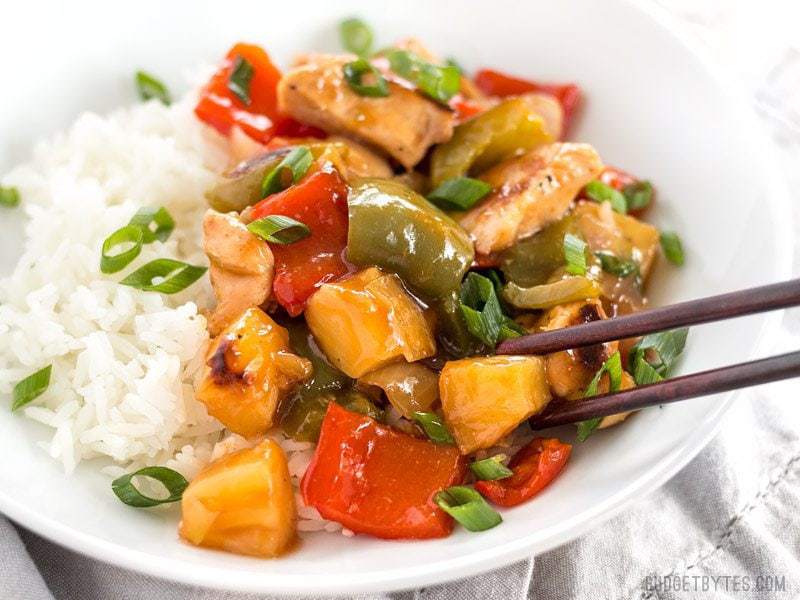 Close up of a plate of Sheet Pan Sweet and Sour Chicken over rice, a piece being picked up with chop sticks