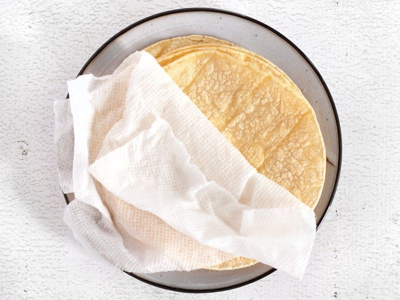 Warm Tortillas stacked on a plate with a damp paper towel on top