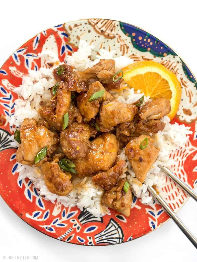 Orange chicken on a bed of rice with an orange slice on a colorful plate. 