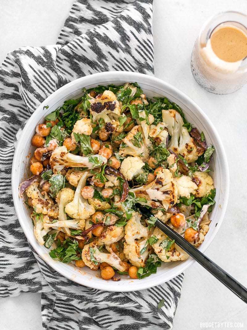 This Roasted Cauliflower Salad combines sweet roasted red onions, spiced chickpeas, tender cauliflower, and a tangy lemon tahini dressing. BudgetBytes.com Overhead shot of a bowl full of Roasted Cauliflower Salad on a striped napkin