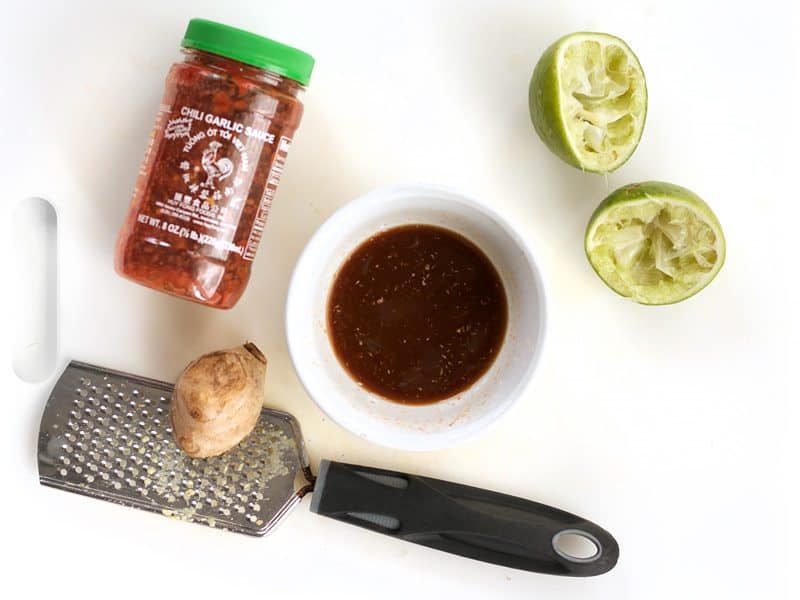 Soy Lime Dressing in a small bowl, surrounded by squeezed limes, chili garlic sauce, and ginger