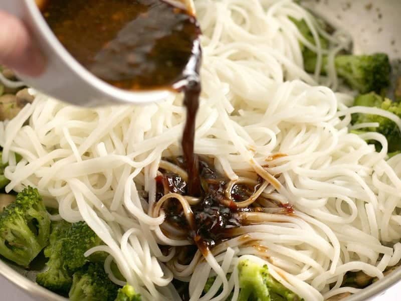 Stir fry sauce being poured over cooked noodles in the skillet with mushrooms and broccoli