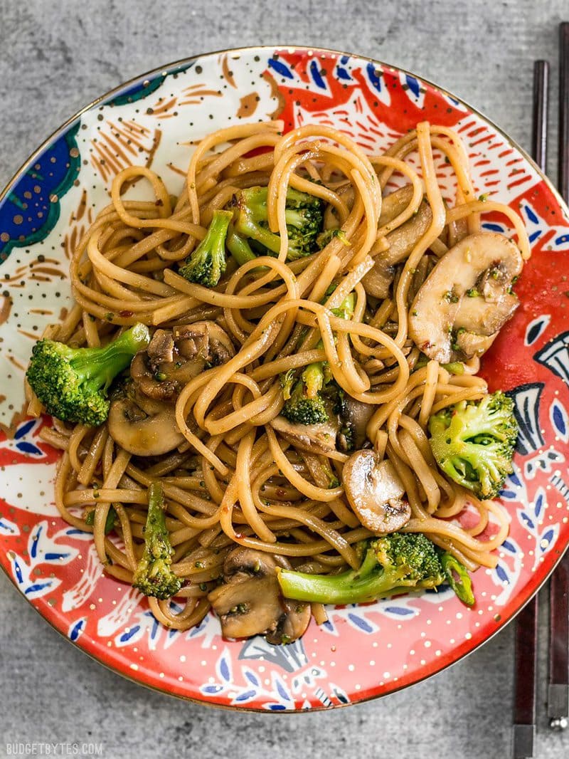 A colorfully patterned plate with Mushroom Broccoli Stir Fry Noodles, chopsticks on the side