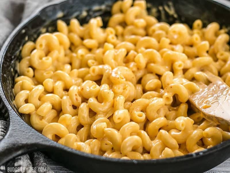 Close up side view of a cast iron skillet full of mac and cheese, with a wooden spoon