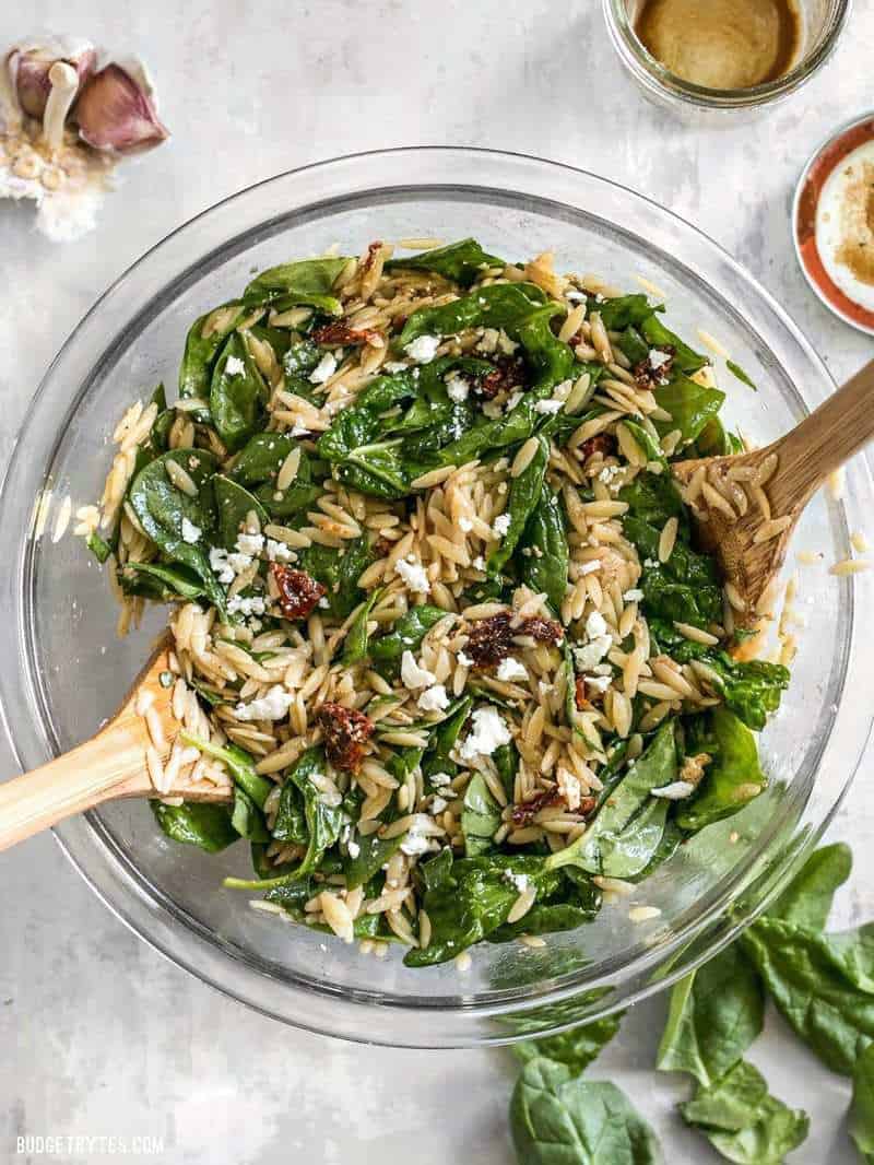 A big glass bowl full of Spinach and Orzo Salad with wooden salad tongs and a jar of dressing on the side